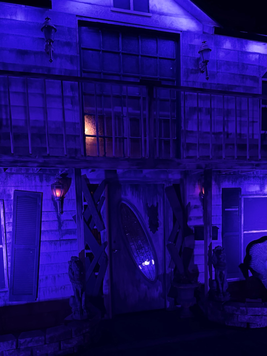 The front of the manor under blue light: boarded windows, stone lions either side of the door, a lamp burning orange on the porch